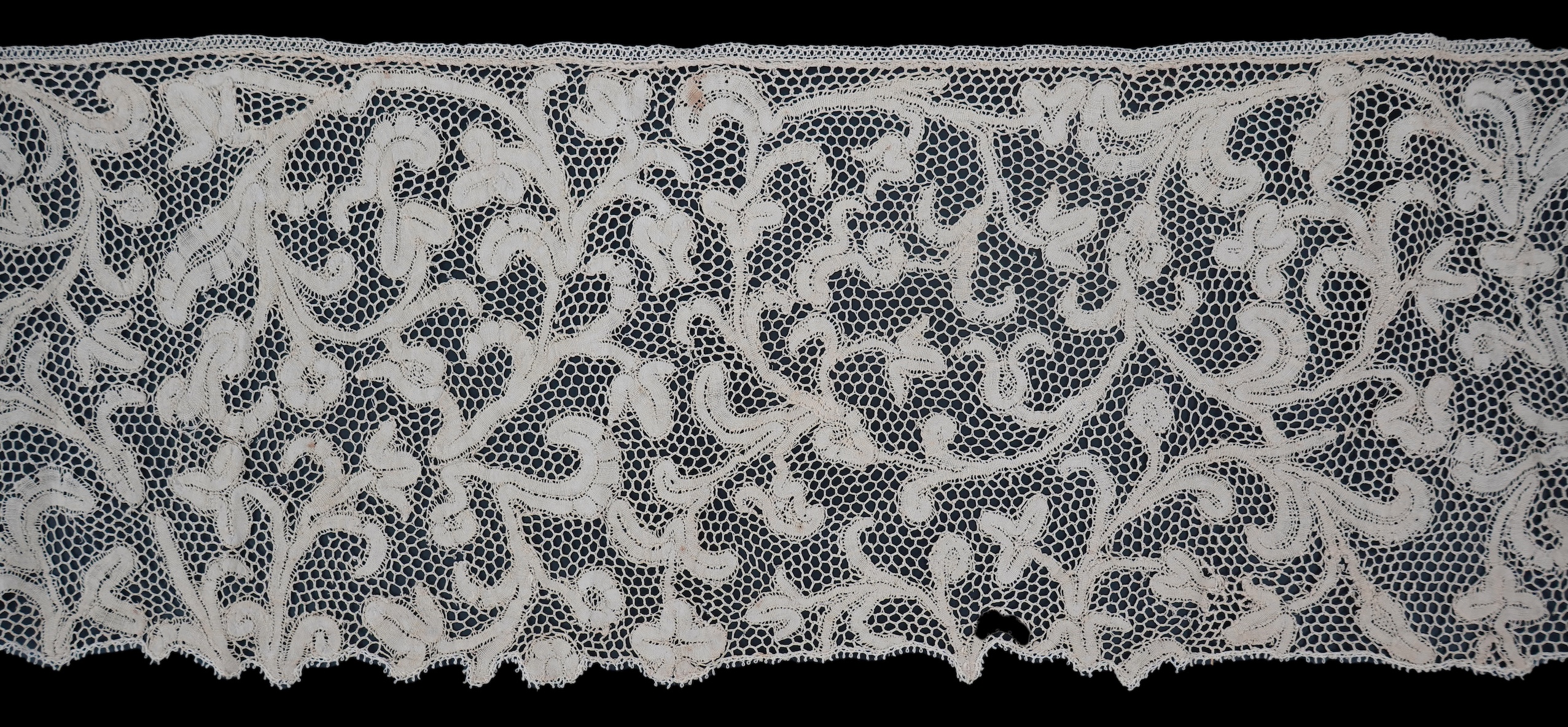 A length of 1740-1760 Italian, Milanese bobbin lace flounce, made from flat braiding or tapes, placed into scrolling patterns, attached to brides, made into a honeycomb mesh. 252cm long x 16cm deep. Condition - one small